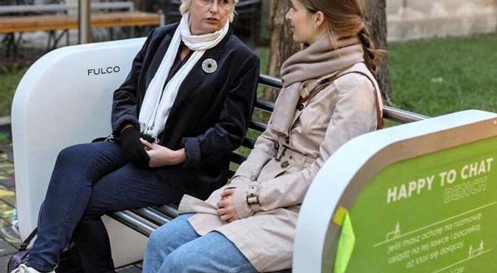 Dois idosos sentados conversando alegremente em um banco de madeira colorido em uma praça ensolarada.