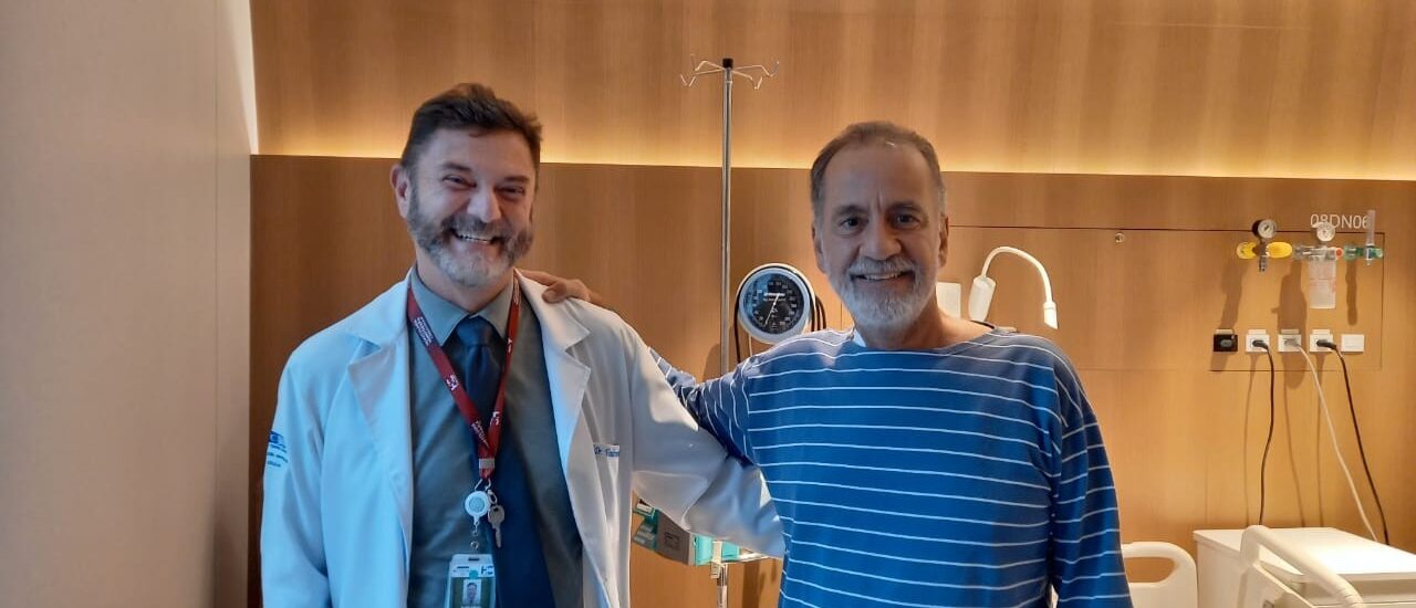 Homem de 61 anos sorrindo ao lado de seu médico em um ambiente hospitalar, celebrando a recuperação de sua saúde.