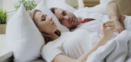 Homem de meia-idade dormindo de lado com semblante desconfortável, enquanto sua parceira ao lado tenta tapar os ouvidos com o travesseiro.